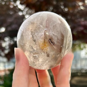 May include: A polished, spherical clear quartz crystal ball featuring internal fractures, cloudy white inclusions, and a distinct golden-yellow mineral deposit near the center, showcasing natural variations and transparency throughout the stone.
