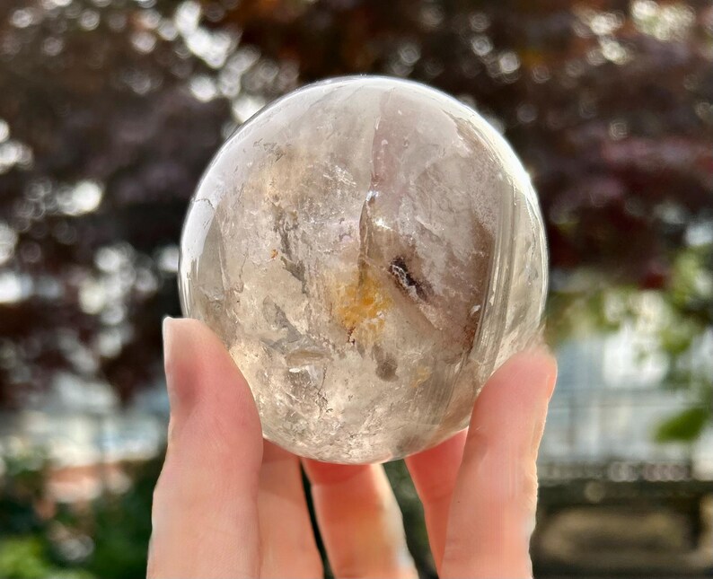 May include: A polished, spherical clear quartz crystal ball featuring internal fractures, cloudy white inclusions, and a distinct golden-yellow mineral deposit near the center, showcasing natural variations and transparency throughout the stone.