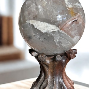 May include: A polished, spherical clear quartz crystal ball featuring internal white and reddish-brown mineral inclusions and fractures. The sphere rests securely on a dark brown, carved wooden stand with a flared base and scalloped top.