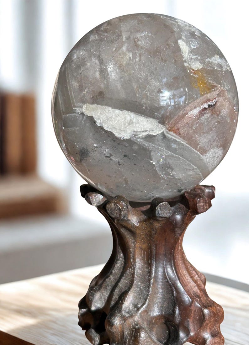 May include: A polished, spherical clear quartz crystal ball featuring internal white and reddish-brown mineral inclusions and fractures. The sphere rests securely on a dark brown, carved wooden stand with a flared base and scalloped top.