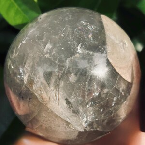 May include: A polished, spherical clear quartz crystal ball featuring internal fractures, cloudy white inclusions, and subtle rainbow light refractions throughout its transparent, glass-like surface, showcasing natural mineral formations.
