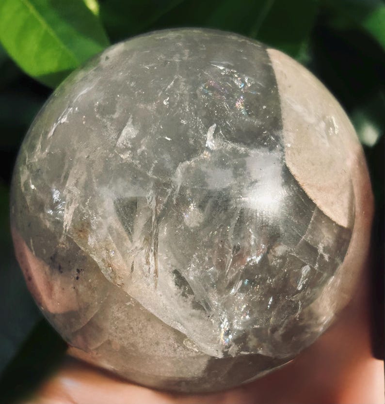 May include: A polished, spherical clear quartz crystal ball featuring internal fractures, cloudy white inclusions, and subtle rainbow light refractions throughout its transparent, glass-like surface, showcasing natural mineral formations.