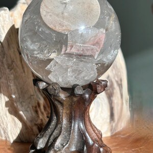 May include: A clear, spherical quartz crystal ball featuring internal mineral inclusions and fractures, resting on a dark, carved wooden stand with a textured, organic shape that mimics the appearance of gnarled tree roots or branches.