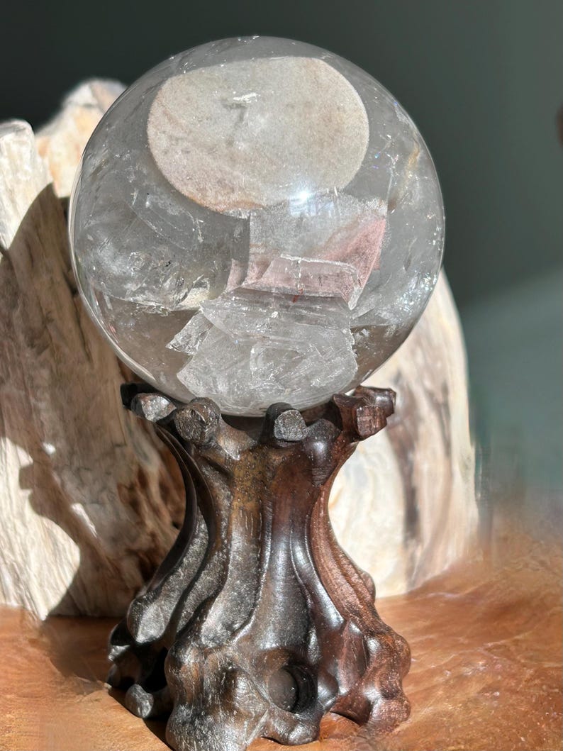 May include: A clear, spherical quartz crystal ball featuring internal mineral inclusions and fractures, resting on a dark, carved wooden stand with a textured, organic shape that mimics the appearance of gnarled tree roots or branches.