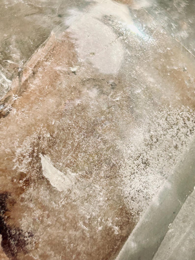 May include: A clear, textured glass surface featuring a mottled, semi-translucent appearance with patches of white, cloudy residue and earthy brown undertones. The material displays an uneven, granular finish with light reflecting off the edges.