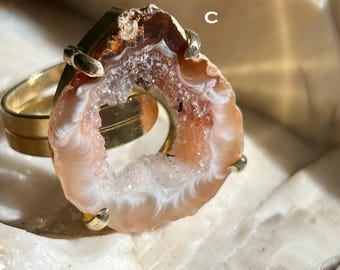 May include: A gold ring with a large, natural agate stone. The agate is a light brown color with white and pink swirls. The ring is set in a gold band.
