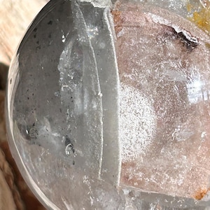 May include: A clear, spherical quartz crystal featuring internal fractures and inclusions. The surface is smooth and reflective, revealing a complex interior with white, grey, and subtle orange mineral deposits trapped within the transparent structure.