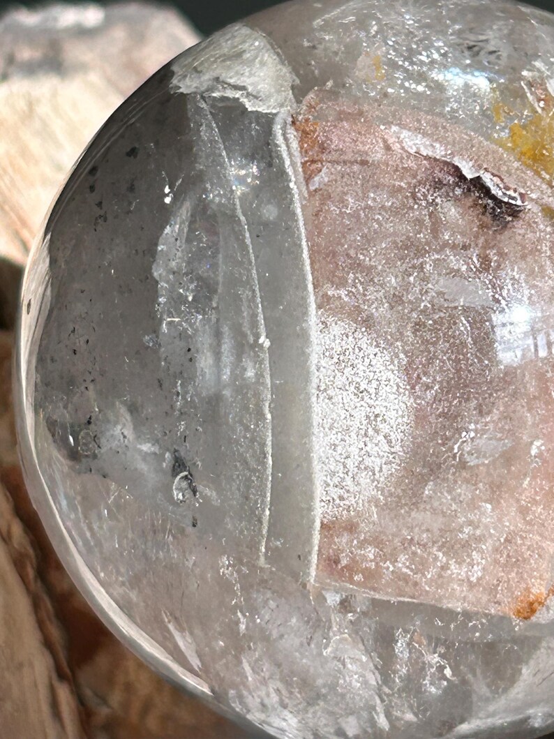May include: A clear, spherical quartz crystal featuring internal fractures and inclusions. The surface is smooth and reflective, revealing a complex interior with white, grey, and subtle orange mineral deposits trapped within the transparent structure.