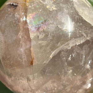May include: A clear, spherical quartz crystal ball featuring internal fractures, natural inclusions, and a distinct iridescent rainbow refraction visible within the translucent stone. The surface is smooth and polished, reflecting light.