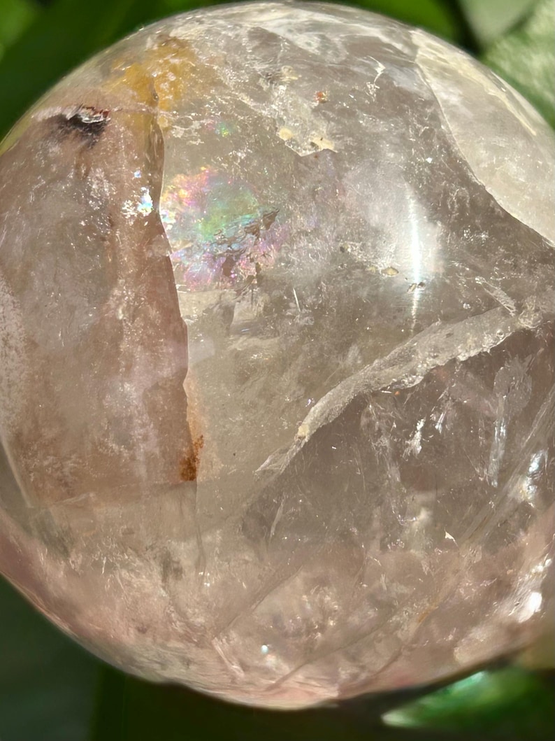 May include: A clear, spherical quartz crystal ball featuring internal fractures, natural inclusions, and a distinct iridescent rainbow refraction visible within the translucent stone. The surface is smooth and polished, reflecting light.