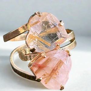 May include: A gold ring with two heart-shaped gemstones. The gemstones are a translucent pink with golden inclusions. The ring has a split shank design, with the gemstones set on top of the band.