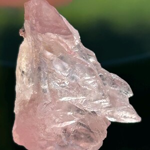 May include: A close-up of a raw rose quartz crystal. The crystal has a translucent, pale pink hue with a rough, textured surface. The light catches the facets, creating a shimmering effect. The background is blurred, highlighting the crystal's details.