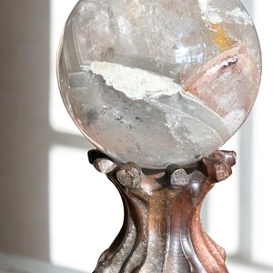 May include: A clear, spherical quartz crystal ball featuring internal white and reddish-brown mineral inclusions, resting securely on a dark, carved wooden stand with a textured, organic shape and a polished finish.