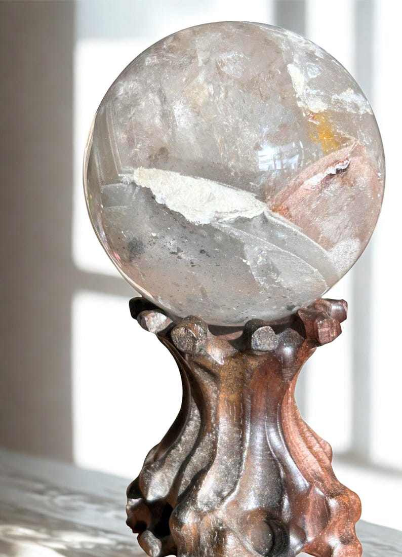 May include: A clear, spherical quartz crystal ball featuring internal white and reddish-brown mineral inclusions, resting securely on a dark, carved wooden stand with a textured, organic shape and a polished finish.