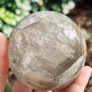 May include: A clear, spherical quartz crystal ball featuring internal fractures, natural inclusions, and visible geometric phantom layers. The surface is smooth and polished, revealing a complex, multi-faceted interior structure within the transparent stone.