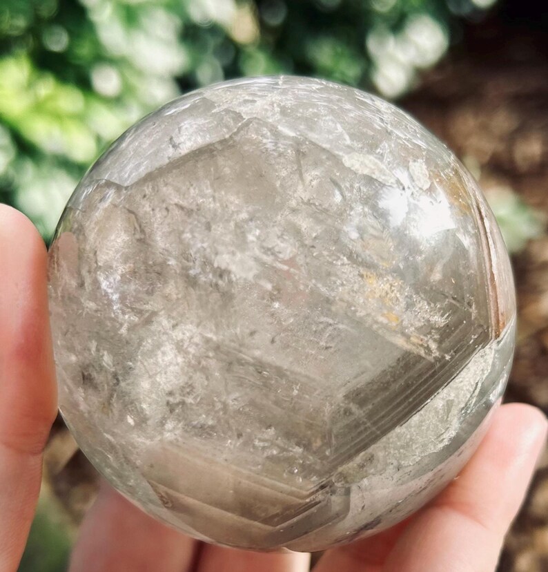 May include: A clear, spherical quartz crystal ball featuring internal fractures, natural inclusions, and visible geometric phantom layers. The surface is smooth and polished, revealing a complex, multi-faceted interior structure within the transparent stone.