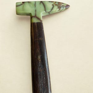 May include: A hammer with a head made of light green stone featuring dark brown veining and a handle crafted from dark brown wood. The head is shaped with a flat striking face on one side and a pointed, tapered end on the opposite side.