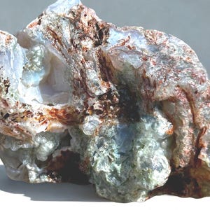 May include: A natural, uncut gemstone with a rough, textured surface. The stone features a mix of colors, including white, brown, and hints of blue and green. The overall shape is irregular, with a crystalline appearance.