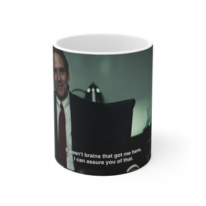 May include: White ceramic mug with a black and white image of a man in a suit and tie. The text on the mug reads "It wasn't brains that got me here, I can assure you of that."