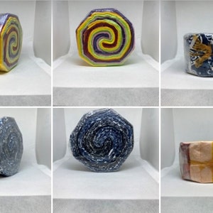 May include: Six fabric rolls, each with a different pattern. The rolls are wrapped in plastic and have a hexagonal shape. The patterns include a rainbow swirl, a floral design, a blue and white pattern, a blue and white pattern with gold accents, a blue and white pattern with black accents, and a yellow and orange pattern with red accents.