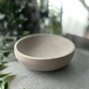 Könnte beinhalten: Eine kleine, runde, hellgraue Schale aus Beton. Die Schale ist glatt und hat einen leicht erhöhten Rand.