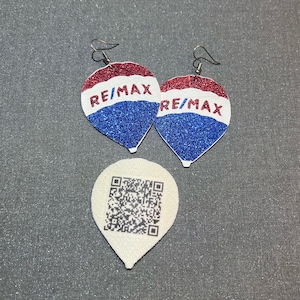 Peut inclure: Boucles d'oreilles scintillantes avec le logo RE/MAX en rouge, blanc et bleu. Les boucles d'oreilles sont en forme de goutte d'eau avec des crochets argentés. Une étiquette blanche en forme de goutte d'eau avec un code QR est en dessous. Sur une surface grise.
