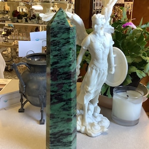 May include: A large green and black crystal point with a faceted shape. The crystal is sitting on a table with a white statue of an angel in the background.