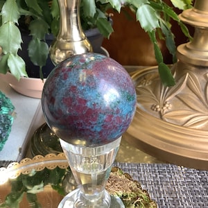 May include: A polished, dark green and red sphere, known as a sphere, sits on a clear glass stand. The sphere is on a gold tray with green moss around the base of the stand.