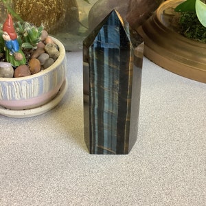 May include: A blue and gold tiger's eye crystal point, also known as a tower or obelisk, is a polished, faceted, and pointed crystal. The crystal is approximately 4 inches tall and has a smooth, polished surface.