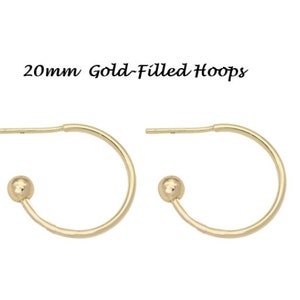 May include: A pair of gold-filled hoop earrings. Each earring has a curved design with a small ball at the bottom and a post for piercing. The text "20mm Gold-Filled Hoops" is above the earrings.