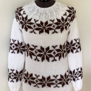 Puede incluir: Jersey blanco de punto con un estampado de copos de nieve marrones. El jersey de manga larga presenta un cuello redondo y un diseño de copos de nieve repetido en el pecho, las mangas y el dobladillo inferior. El jersey se muestra en un maniquí.