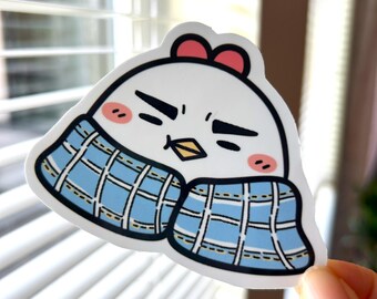 May include: A white sticker featuring a cartoon chicken with a red bow, rosy cheeks, and a blue plaid scarf. The chicken has a grumpy expression. The sticker is held by a hand in front of a window.