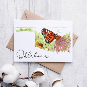 K&ouml;nnte beinhalten: Eine wei&szlig;e Karte mit einer Aquarell-Illustration des Bundesstaates Oklahoma. Die Illustration zeigt einen Monarchfalter, Wildblumen und das Wort "Oklahoma" in Kursivschrift.