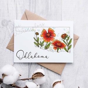 K&ouml;nnte beinhalten: Eine Gru&szlig;karte mit einer Aquarell-Illustration des Bundesstaates Oklahoma mit orangefarbenen und gelben Blumen. Der Text "Oklahoma" ist in Kursivschrift unter der Illustration geschrieben.