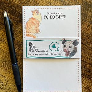 May include: A white tear-away notepad with a dashed orange border, featuring an illustration of an orange tabby cat and the text (do not want) TO DO LIST. A paper band displays the text Okie Watercolors, tear away notepad --50 pages, and a possum.