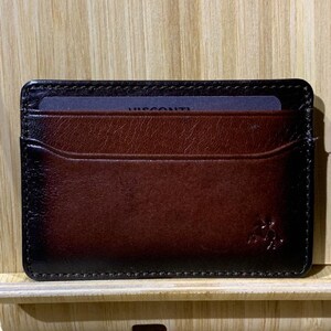 May include: A brown leather card holder with a black stitched border. The card holder has a single card slot and a small pocket on the inside. The Visconti logo is embossed on the front.