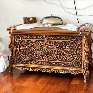 Puede incluir: Un gran cofre de madera tallado intrincadamente con un acabado marrón oscuro. El cofre tiene dos puertas con diseños florales ornamentados y una cerradura en el centro. El cofre está sentado en un suelo de madera.