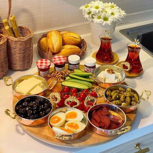 May include: A wooden tray with copper bowls filled with various breakfast items, including olives, eggs, cheese, and cured meat. The tray is surrounded by jars of jam, Nutella, and walnuts. There are also two glasses of red tea and a bouquet of white daisies in the background.