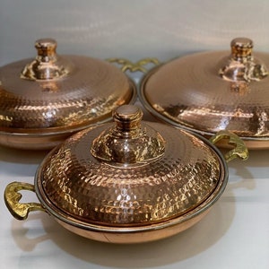 Könnte beinhalten: Drei Servierplatten aus Kupfer mit gehämmerter Oberfläche und goldenen Griffen. Die Platten sind ineinander verschachtelt, wobei die größte Platte unten und die kleinste Platte oben liegt.
