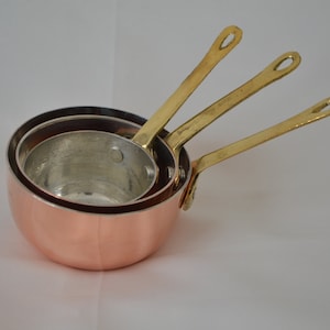 Może przedstawiać: Zestaw trzech miedzianych garnków z mosiężnymi uchwytami. Garnki są włożone jeden w drugi, przy czym największy garnek znajduje się na dole.