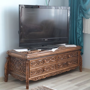 Puede incluir: Un mueble de TV de madera oscura con tallas intrincadas y cuatro cajones. El mueble est&aacute; en una sala de estar con una pared azul claro y una alfombra en el suelo.