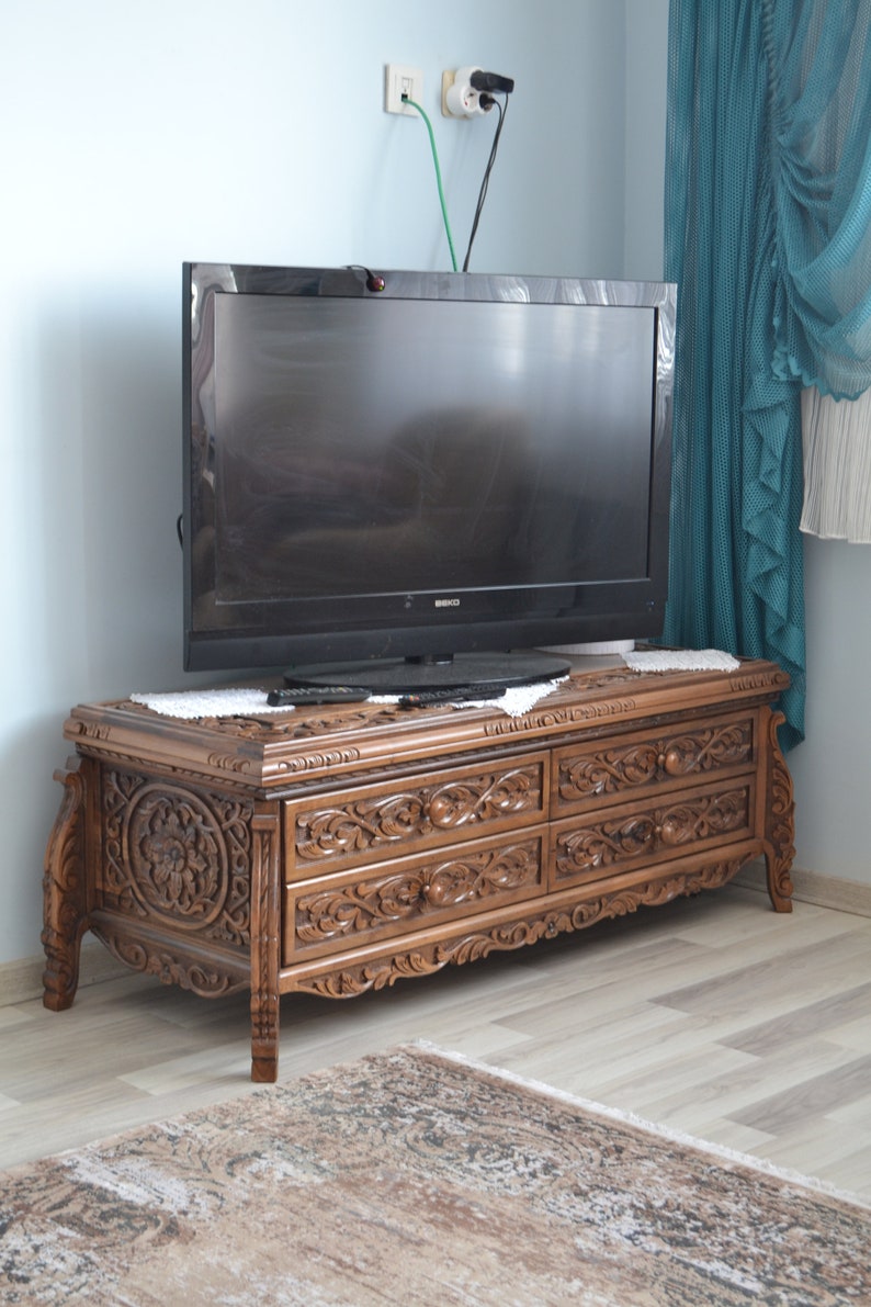 Puede incluir: Un mueble de TV de madera oscura con tallas intrincadas y cuatro cajones. El mueble est&aacute; en una sala de estar con una pared azul claro y una alfombra en el suelo.