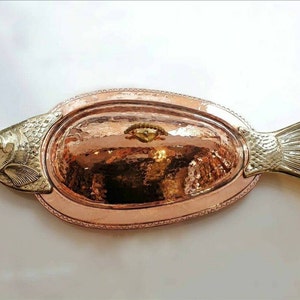 Puede incluir: Una fuente de servir de cobre con forma de pez con acabado martillado. La fuente tiene una sección ovalada elevada en el centro y detalles de cabeza y cola de pez en tono dorado.