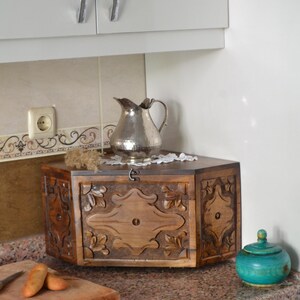 May include: A wooden bread box with carved floral designs, a silver pitcher, and a small turquoise jar sit on a granite countertop. The bread box has a hinged door and a decorative lock. The kitchen setting includes white cabinets.