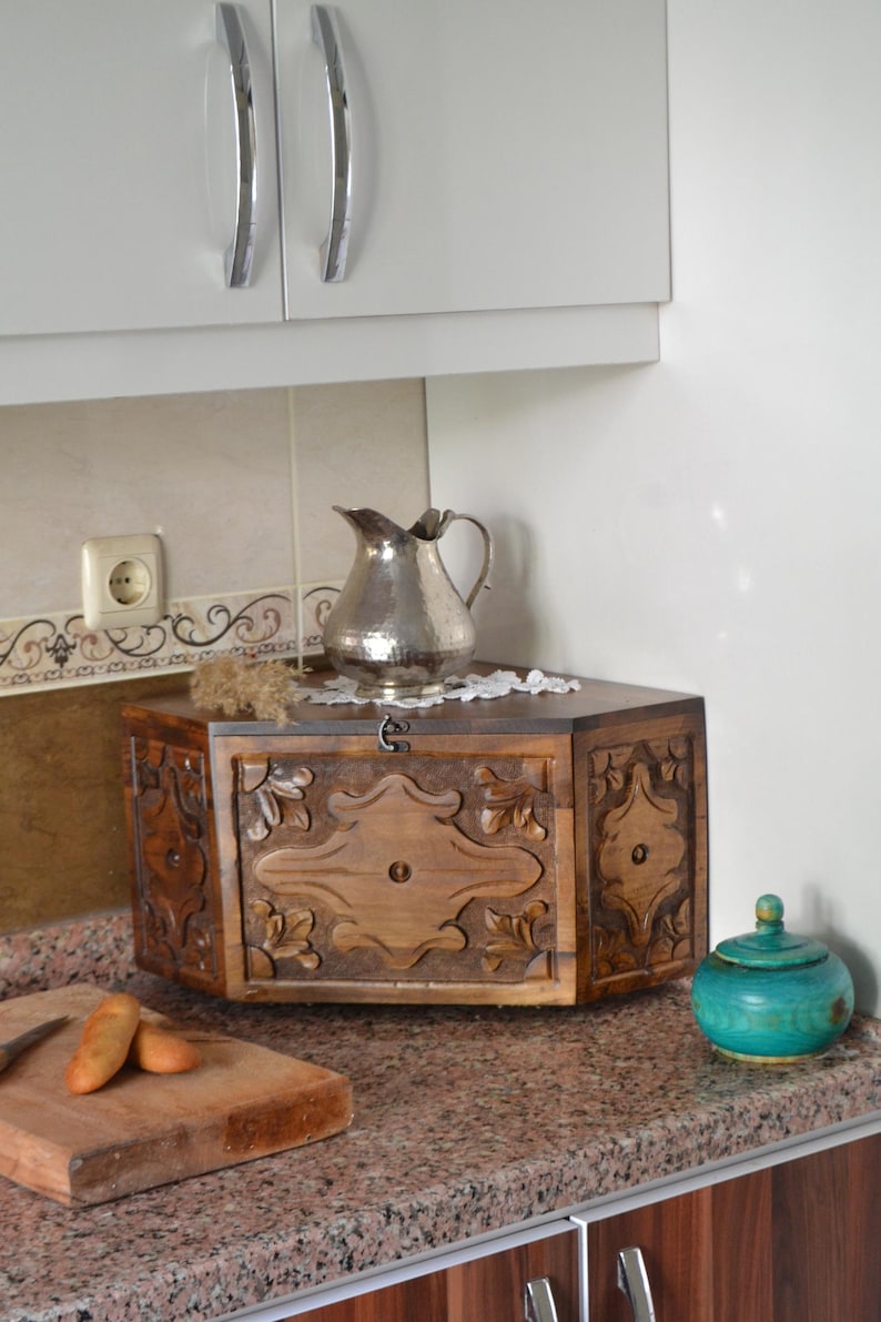 May include: A wooden bread box with carved floral designs, a silver pitcher, and a small turquoise jar sit on a granite countertop. The bread box has a hinged door and a decorative lock. The kitchen setting includes white cabinets.