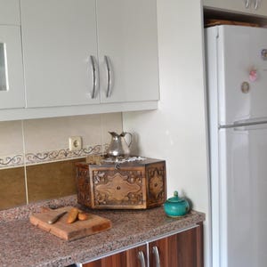 May include: A kitchen scene featuring a wooden cutting board with carrots and a knife, a decorative wooden box, and a small teal jar. The countertop has a speckled granite pattern, and the cabinets are white and wood-toned.