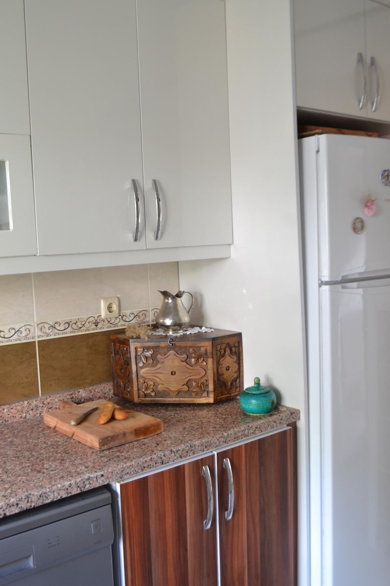 May include: A kitchen scene featuring a wooden cutting board with carrots and a knife, a decorative wooden box, and a small teal jar. The countertop has a speckled granite pattern, and the cabinets are white and wood-toned.