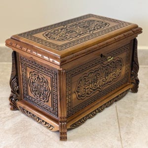 Puede incluir: Un cofre de madera con intrincadas tallas florales. El cofre es de color marrón cálido con acentos más oscuros en los detalles tallados. Presenta una forma rectangular con una tapa con bisagras y una pequeña cerradura. El cofre está sostenido por patas cortas y decorativas.