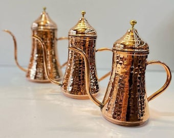 May include: Three copper coffee pots with hammered texture and brass accents. The pots have a rounded body and a spout on the side.