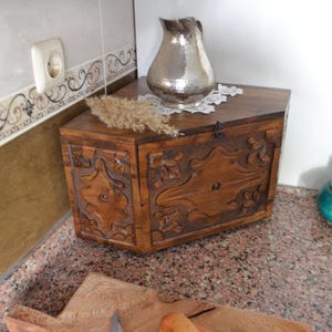 May include: A wooden chest with carved floral designs, a silver pitcher, and a small turquoise jar with a lid. The chest is brown, and the pitcher has a hammered texture. A sprig of dried grass sits on top of the chest.